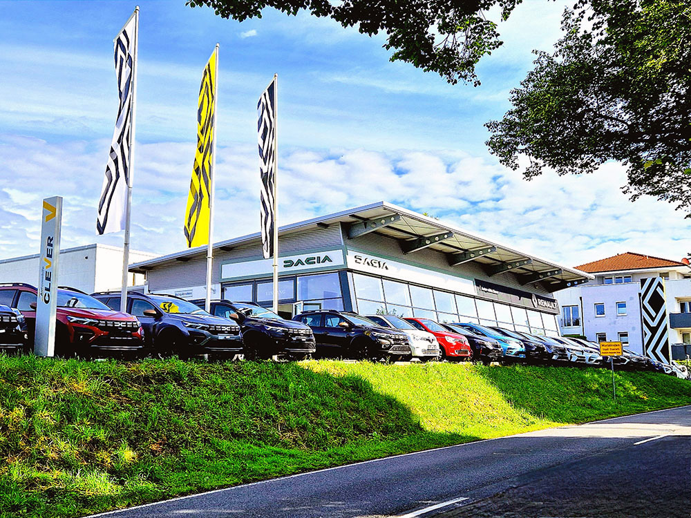 Automobile Clever Werkstatt Das Autohaus Clever mit einer Reihe geparkter Fahrzeuge, darunter mehrere Renault-Modelle, vor einem modernen Verkaufsgebäude mit Fahnen und dem Renault-Logo.